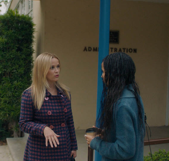 A woman wearing a plaid coat and a red pin is talking to another woman. Description generated by AI